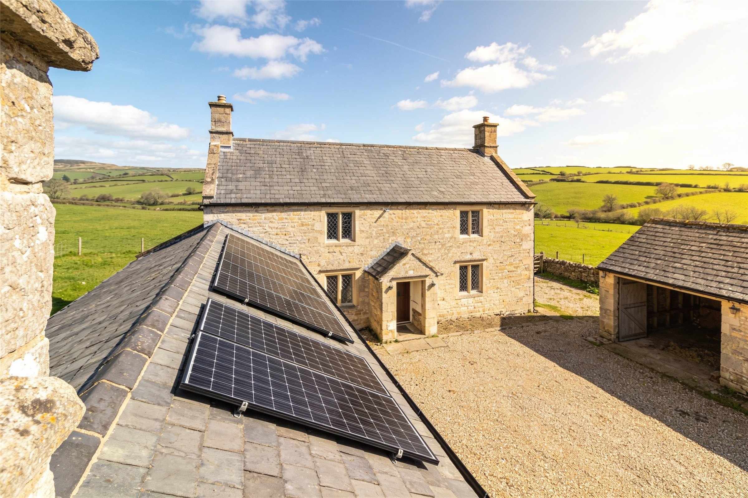 The same stone farmhouse in bright sun, two neat rows of black solar panels low on its long slate roof, fields and white clouds beyond