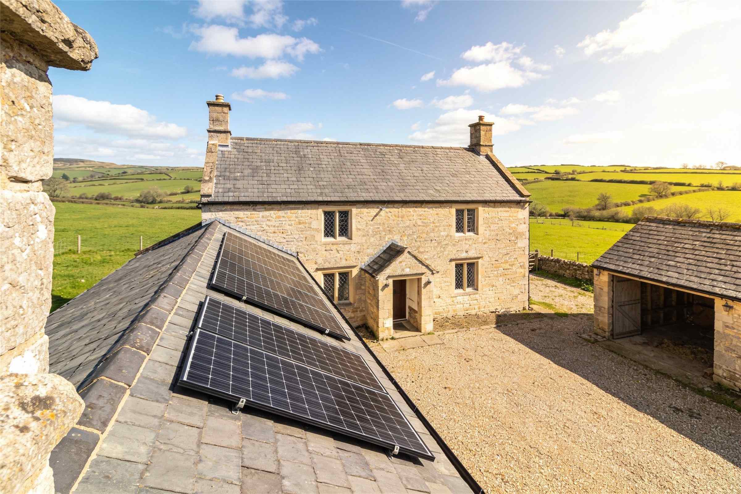 The same stone farmhouse in bright sun, two neat rows of black solar panels low on its long slate roof, fields and white clouds beyond