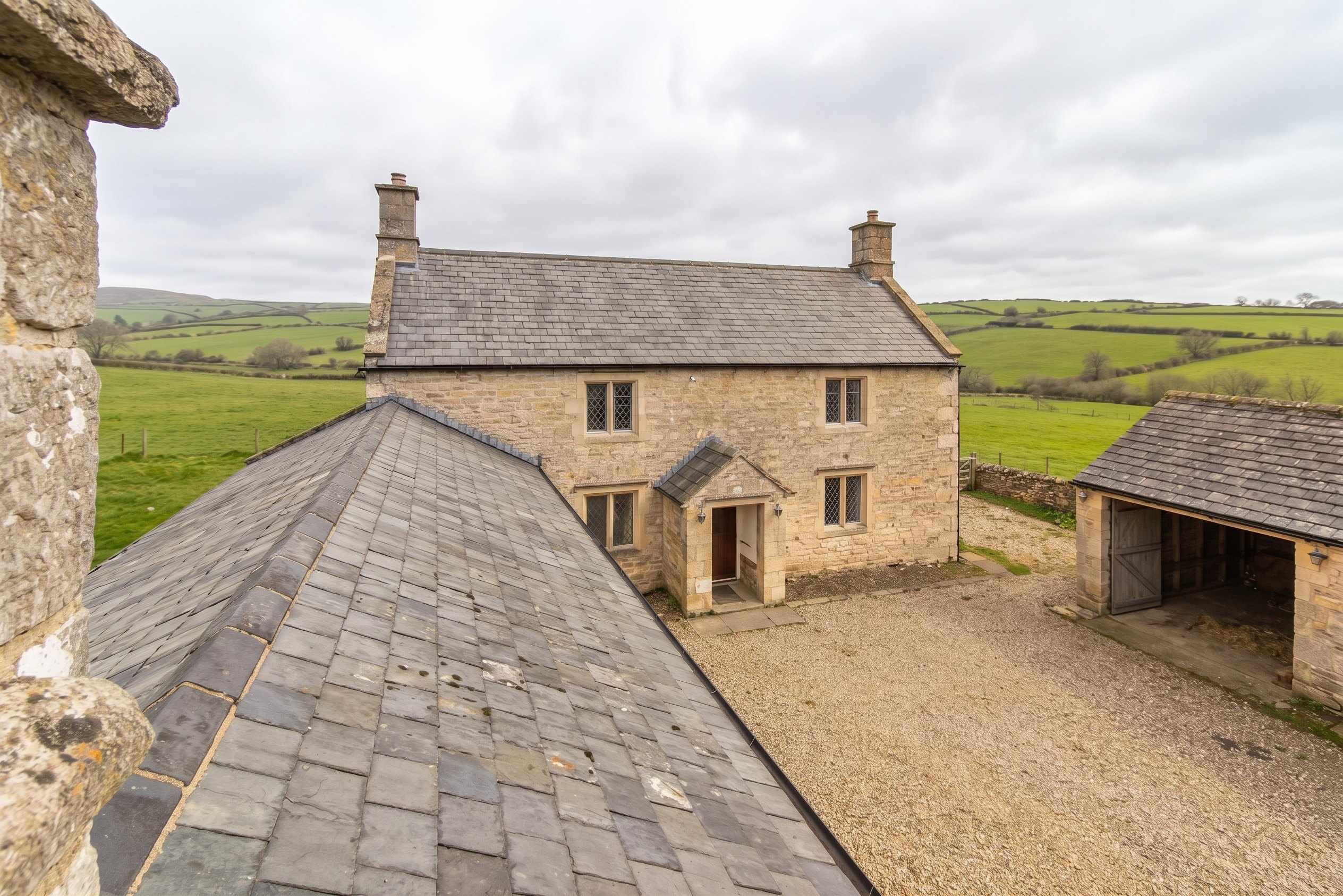 An old stone farmhouse with a long plain slate roof on a grey morning, seen across a gravel farmyard with fields beyond